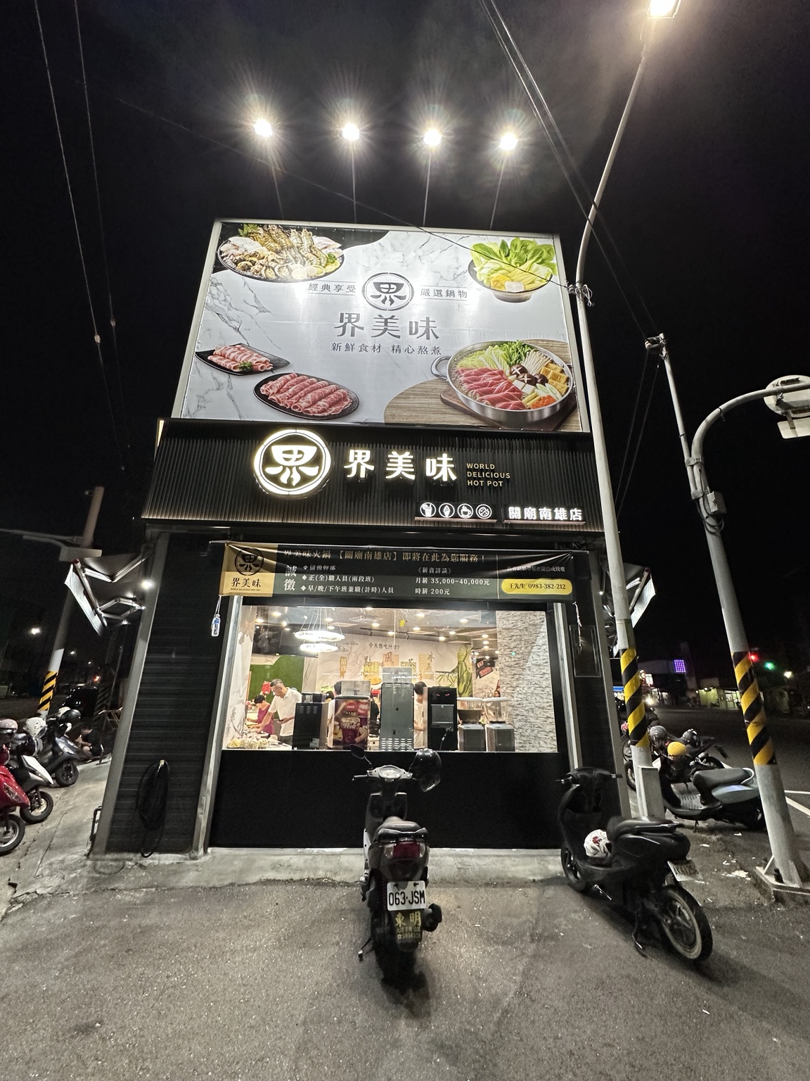 界美味關廟南雄店正面夜景，投射燈照亮大型帆布與下方店名招牌